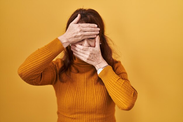 Hispanische Frau mittleren Alters, die vor gelbem Hintergrund steht und Augen und Mund mit den Händen bedeckt, ist überrascht und schockiert und verbirgt Emotionen