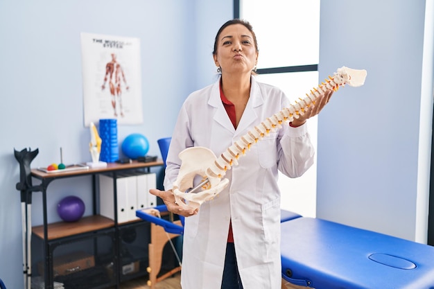 Hispanische Frau mittleren Alters hält ein anatomisches Modell der Wirbelsäule in der Hand und blickt in die Kamera, während sie einen Kuss zuwirft, was ein schöner und sexy Liebesausdruck ist