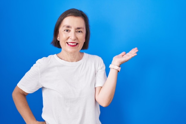 Hispanische Frau mittleren Alters steht vor blauem Hintergrund, lächelt fröhlich, präsentiert und zeigt mit der Handfläche in die Kamera.