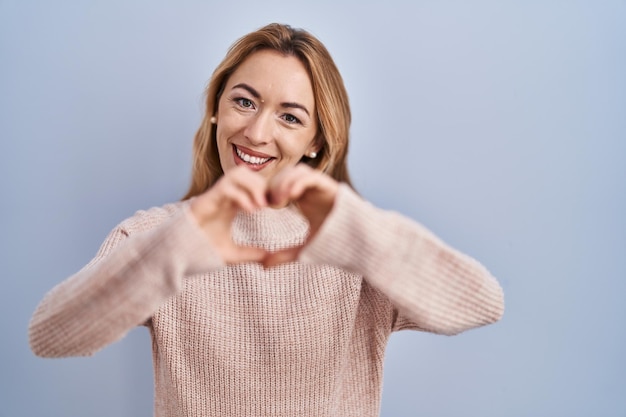 Hispanische Frau steht vor blauem Hintergrund und lächelt verliebt und macht mit den Händen ein romantisches Konzept in Form eines Herzsymbols