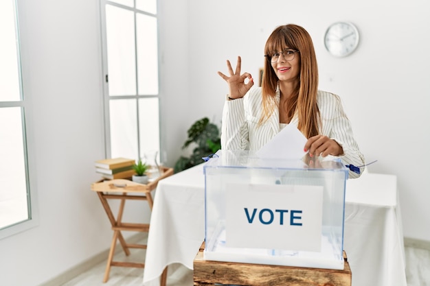 Hispanische Geschäftsfrau, die zur Wahl geht und einen Umschlag in die Wahlurne steckt, lächelt positiv und macht mit Hand und Fingern ein gutes Zeichen mit erfolgreichem Gesichtsausdruck
