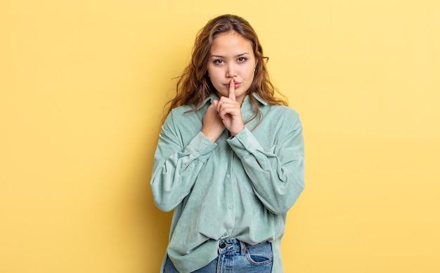 Hispanische hübsche Frau, die ernst und kreuzig aussieht, mit dem Finger auf die Lippen gedrückt, die Schweigen fordert oder ein Geheimnis bewahrt