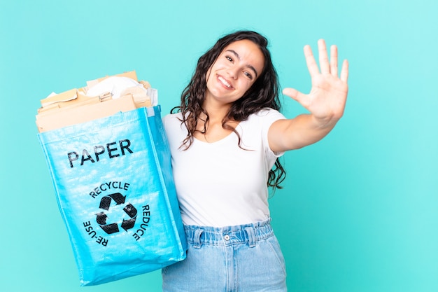 Hispanische hübsche Frau, die lächelt und freundlich aussieht, Nummer fünf zeigt und eine Recyclingpapiertüte hält