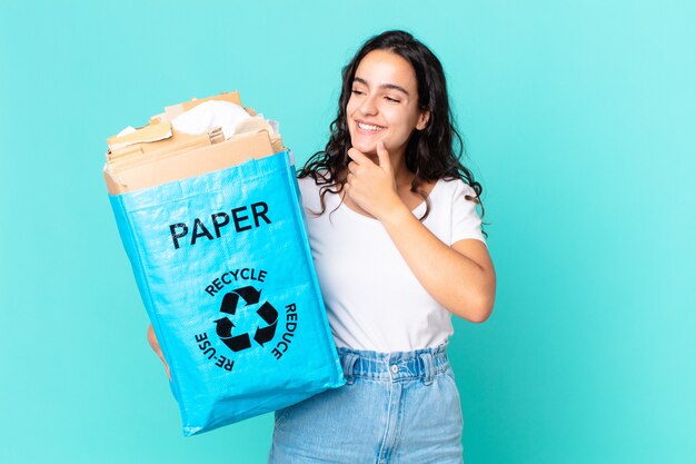 Hispanische hübsche Frau, die mit einem glücklichen, selbstbewussten Ausdruck mit der Hand am Kinn lächelt und eine Recyclingpapiertüte hält