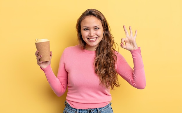 Hispanische hübsche Frau, die sich glücklich fühlt, wenn sie Zustimmung mit einer okay-Geste zum Mitnehmen des Kaffeekonzepts zeigt