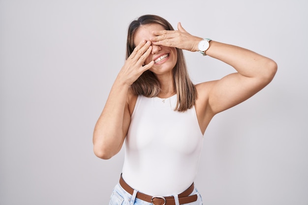 Hispanische junge Frau, die über weißem Hintergrund steht und die Augen mit Händen bedeckt, die fröhlich und lustig lächeln. blindes Konzept.