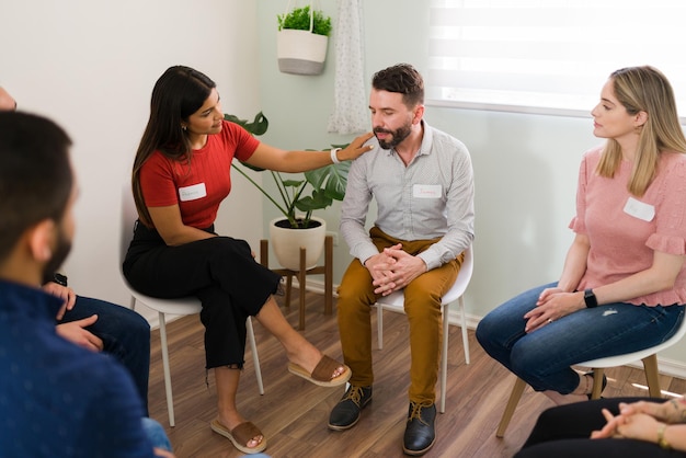 Hispanische junge Frau tröstet ein depressives männliches Mitglied ihrer Selbsthilfegruppe. Junge Leute beraten einen Mann während eines Aggressionsmanagement-Meetings