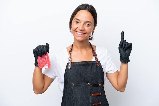 Hispanische Metzgerin, die eine Schürze trägt und frisch geschnittenes Fleisch serviert, isoliert auf weißem Hintergrund, zeigt und hebt einen Finger als Zeichen der Besten