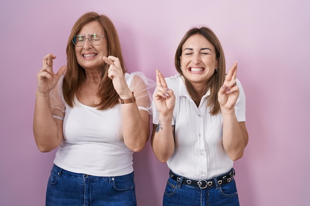 Hispanische Mutter und Tochter tragen ein beiläufiges weißes T-Shirt auf einem rosa Hintergrund, gestalten mit gekreuzten Fingern, lächeln mit Hoffnung und geschlossenen Augen. Glück und abergläubisches Konzept