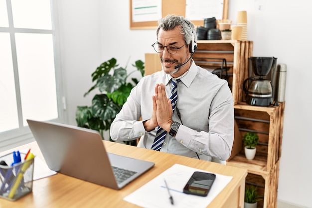 Hispanischer Geschäftsmann mittleren Alters, der im Büro arbeitet und ein Operator-Headset trägt, das mit den Händen zusammen betet und um Vergebung bittet und selbstbewusst lächelt