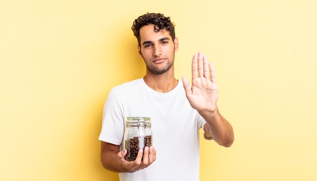 Hispanischer gutaussehender Mann, der ernst aussieht und eine offene Handfläche zeigt, die eine Stopp-Geste macht. Kaffeebohnen Flasche