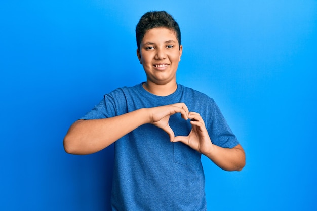 Hispanischer Junge des Teenagers, der lässiges blaues T-Shirt trägt, das in der Liebe lächelt, die Herzsymbolform mit den Händen tut. romantisches Konzept.