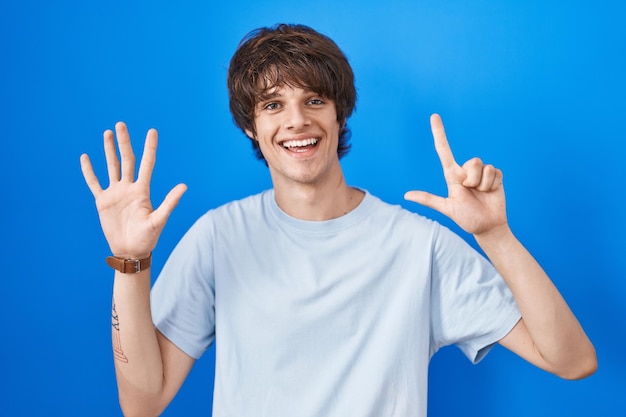 Hispanischer junger Mann, der vor blauem Hintergrund steht und mit den Fingern Nummer sieben nach oben zeigt, während er selbstbewusst und glücklich lächelt.