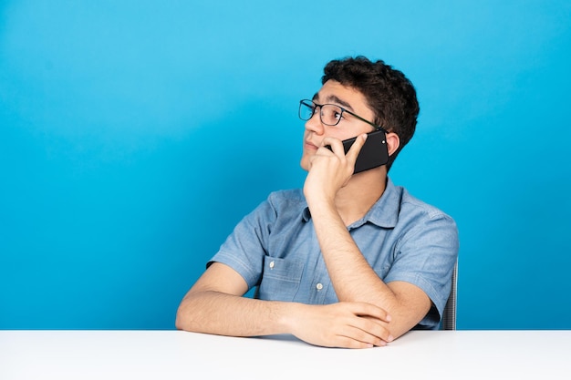 Hispanischer junger Mann sitzt und telefoniert isoliert auf blauem Hintergrund