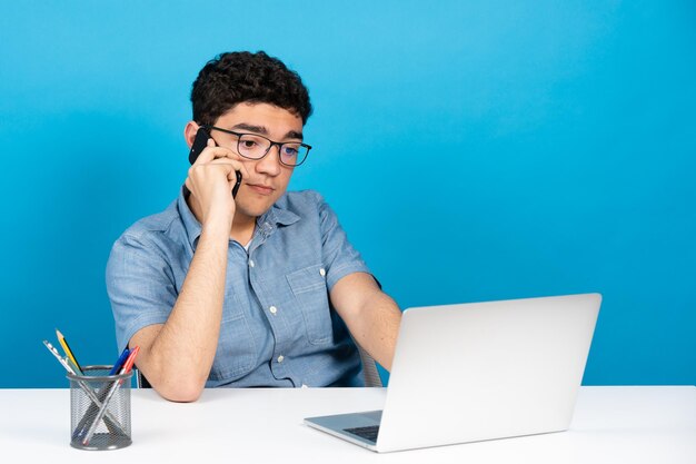 Hispanischer junger Mann telefoniert vor Laptop isoliert auf blauem Hintergrund