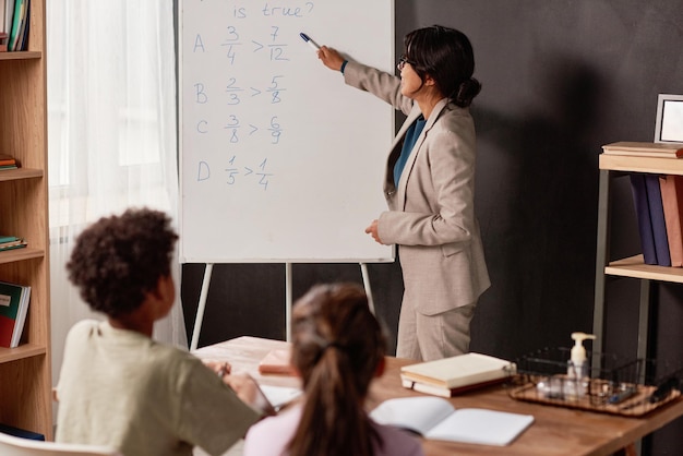 Hispanischer Lehrer mit Haarknoten, der auf Whiteboard zeigt, während er mit Kindern im Klassenzimmer arbeitet