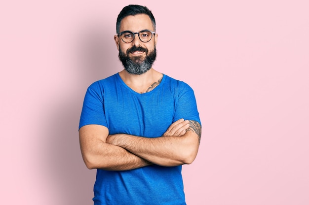Hispanischer Mann mit Bart, legerem T-Shirt und Brille, glückliches Gesicht, lächelnd und mit verschränkten Armen, der positiv in die Kamera blickt