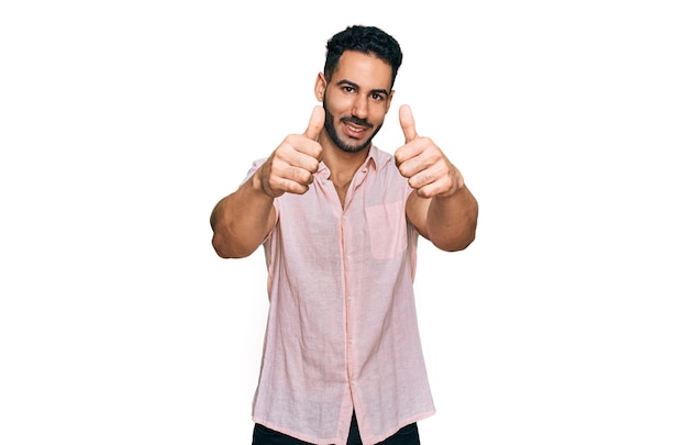 Hispanischer Mann mit Bart und lässigem Hemd, der zustimmend eine positive Geste macht, mit erhobenem Daumen, lächelnd und glücklich über den Erfolg, Siegergeste