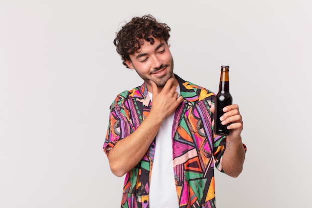 Hispanischer Mann mit Bier, der mit einem glücklichen, selbstbewussten Ausdruck mit der Hand am Kinn lächelt, sich wundert und zur Seite schaut
