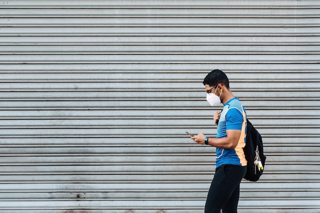 Hispanischer Mann mit einer schützenden Gesichtsmaske und einem Rucksack, der sein Telefon vor einer Garage anschaut