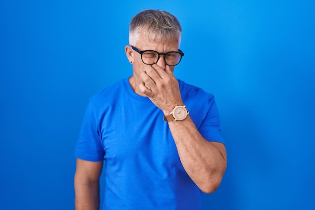 Hispanischer Mann mit grauen Haaren, der vor blauem Hintergrund steht und etwas stinkenden und ekelhaften, unerträglichen Geruch riecht und den Atem anhält, die Finger auf der Nase, schlechter Geruch