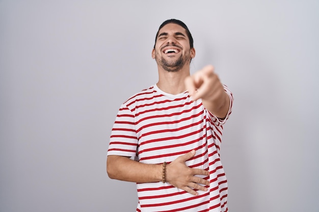 Hispanischer Mann mit langen Haaren, der vor isoliertem Hintergrund steht und Sie auslacht, mit dem Finger in die Kamera zeigt und die Hand über dem Körper beschämt