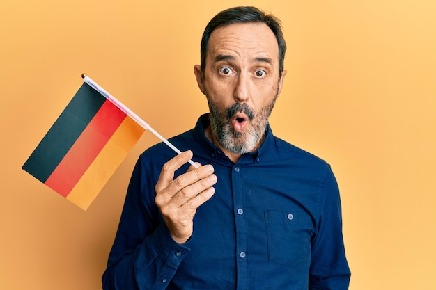 Hispanischer mann mittleren alters, der deutschland-flagge hält, erschrocken und erstaunt mit offenem mund für überraschendes ungläubiges gesicht