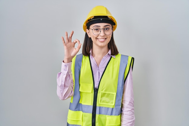 Hispanisches Mädchen in Bauarbeiteruniform und Bauarbeiterhelm lächelt positiv und macht mit Hand und Fingern ein „OK“-Zeichen mit erfolgreichem Gesichtsausdruck