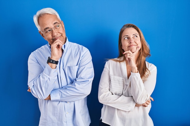 Hispanisches Paar mittleren Alters, das über blauem Hintergrund steht, mit der Hand am Kinn, das über Fragen nachdenkt, nachdenklicher Ausdruck. lächelndes und nachdenkliches Gesicht. Zweifel Konzept.