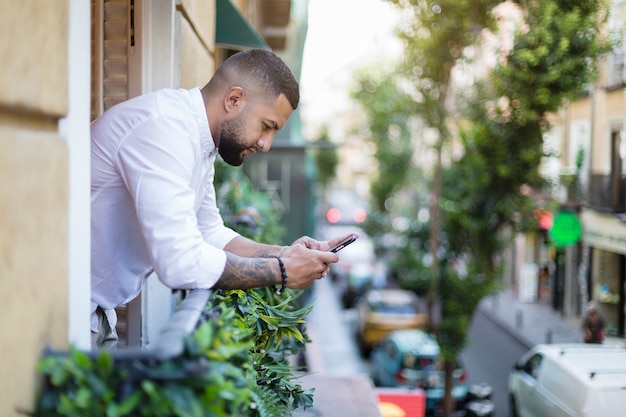 Hispano de sexo masculino adulto medio que tiene un momento de descanso. Está usando su teléfono inteligente asomado a la ventana. Espacio para texto.