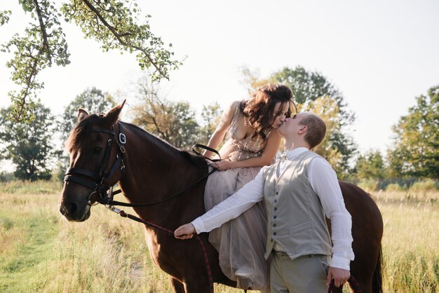 Historia de amor y boda cerca del río en un campo al atardecer con caballo marrón. La novia en vestido aireado es del color de la rosa polvorienta. Vestido beige con destellos. la novia y el novio se abrazan y besan.