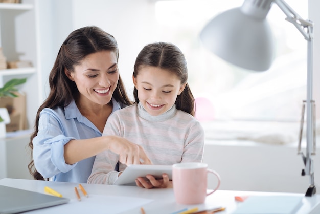 Historia interesante. Linda chica positiva positiva sonriendo y leyendo un libro mientras está sentado junto con su madre