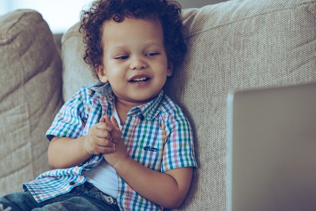 Historieta divertida. Pequeño bebé africano alegre que mira su computadora portátil con una sonrisa mientras está sentado en el sofá en casa