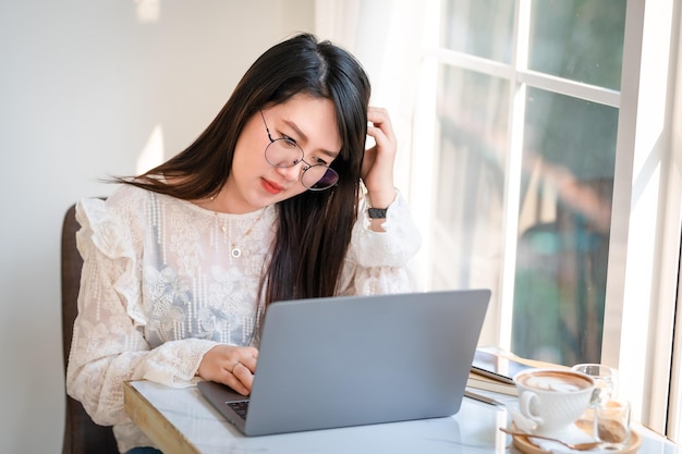 Hizo hincapié en la gente independiente de negocios mujer asiática que tiene dolor de cabeza mientras trabaja usando una computadora portátil con una taza de café y un teléfono inteligente en la ventana de la cafetería como el fondo