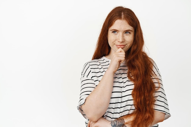 Hmm interessante. Mulher jovem ruiva com cabelo longo natural pensando, sorrindo satisfeito e desviando o olhar para o banner de venda, tem uma ideia, um bom plano, ponderar smth, de pé contra um fundo branco