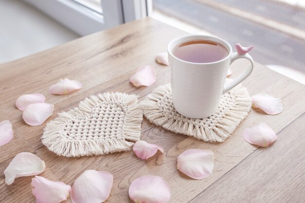 Hobby artesanal de macramé. Chá em uma xícara na montanha-russa de macramê branca na mesa de madeira com pétalas de rosa. Estilista de alimentos. Eco macramé decoração para casa. Dia de São Valentim