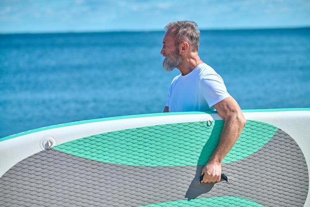 Hobby. Hombre confiado de cabello gris barbudo de lado a la cámara sosteniendo una tabla de remo contra el mar azul y el cielo en un día soleado