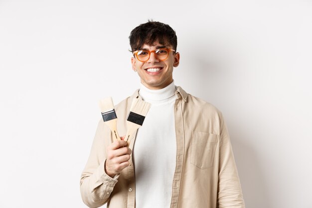Hobbys und Freizeitkonzept. Stilvoller junger Mann mit Brille, der Pinsel zeigt und lächelt und einlädt, etwas zu malen und auf weißem Hintergrund steht.