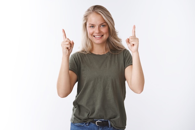 Hoch gehen wir. Schöne europäische Frau mit blonden Haaren in olivgrünem T-Shirt, die die Zeigefinger nach oben hebt und versichert lächelt, den perfekten Ort zu empfehlen, der Ratschläge gibt, welches Produkt wählen?