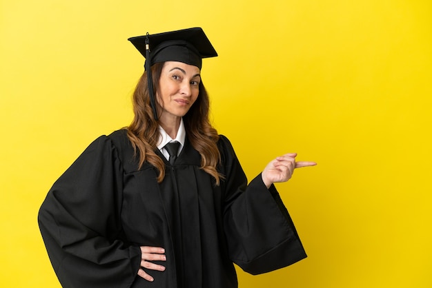 Hochschulabsolvent mittleren Alters isoliert auf gelbem Hintergrund, der mit dem Finger zur Seite zeigt und ein Produkt präsentiert