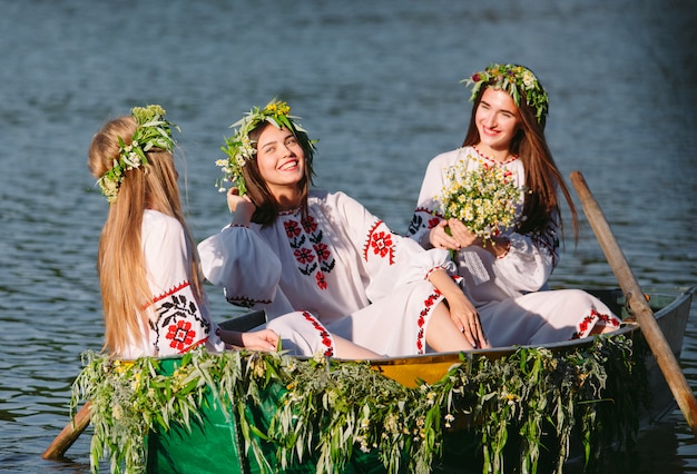 Hochsommer. Junge Mädchen in Trachten segeln in einem Boot, das mit Blättern und Pflanzen geschmückt ist. Slawischer Feiertag von Ivan Kupala.