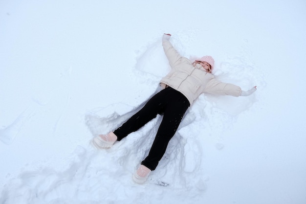 Hochwinkelansicht einer glücklichen Frau, die auf Schnee liegt und ihre Arme und Beine auf und ab bewegt, wodurch eine Schneeengelfigur entsteht Lächelnde Frau, die im Winterurlaub auf Schnee liegt