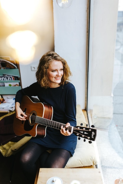 Hochwinkelansicht einer lächelnden jungen Frau, die zu Hause am Fenster Gitarre spielt