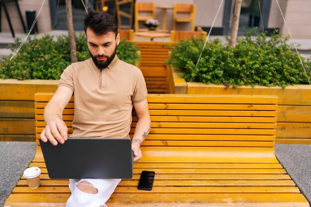 Hochwinkelansicht eines fokussierten bärtigen jungen Mannes, der an einem Laptop arbeitet, der auf einer Bank auf der Stadtstraße mit Kaffee zum Mitnehmen sitzt