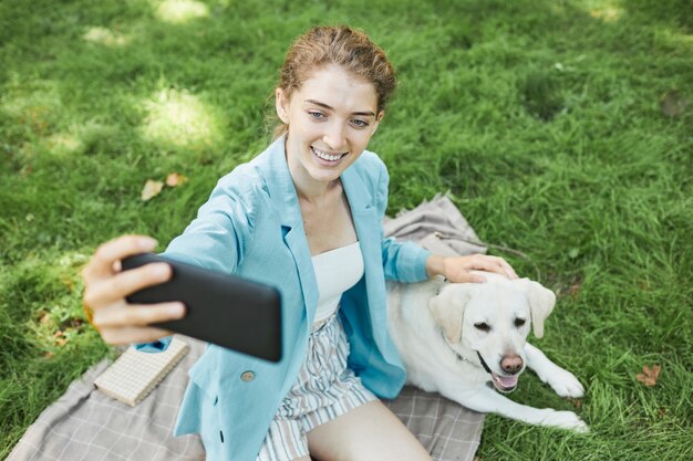 Hochwinkeliges Porträt einer lächelnden jungen Frau, die im Freien ein Selfie mit Hund macht, während sie gemeinsam im Park spazieren geht, Platz kopieren