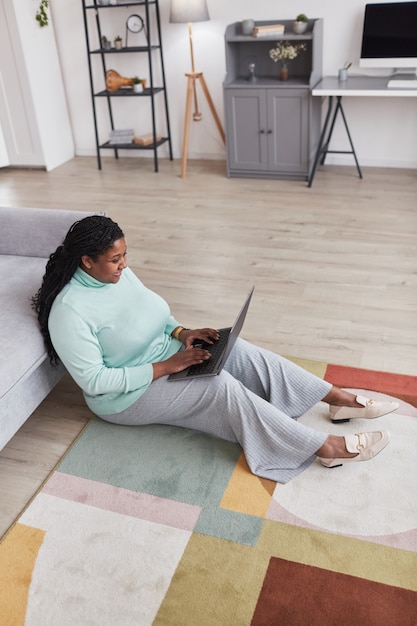 Hochwinkeliges Porträt in voller Länge einer kurvigen Afroamerikanerin, die glücklich lächelt und einen Laptop benutzt, während sie die Arbeit von zu Hause aus auf dem Boden genießt