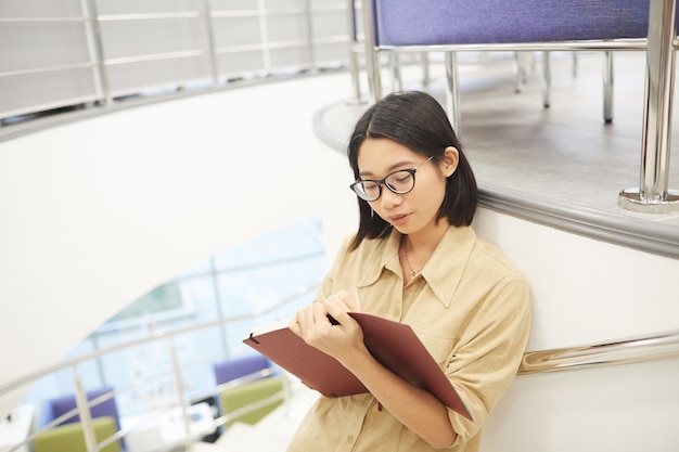 Hochwinkelporträt der jungen asiatischen Frau, die im Notizbuch schreibt