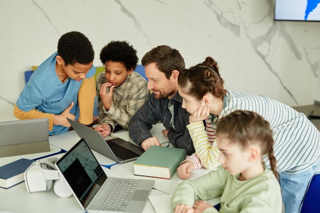 Hochwinkelporträt eines lächelnden männlichen Lehrers mit einer vielfältigen Gruppe von Kindern, die gemeinsam einen Laptop verwenden