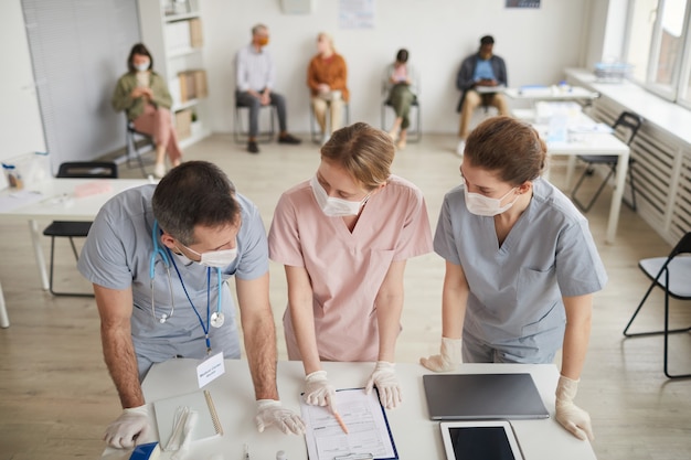 Hochwinkelporträt von drei Ärzten, die Masken tragen, die in der medizinischen Klinik am Schreibtisch stehen, Kopierraum