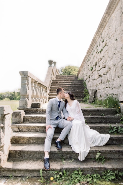 Hochzeit, asiatischer Bräutigam und Braut, die Hände halten, während sie auf der alten Steintreppe neben altem Schloss sitzen